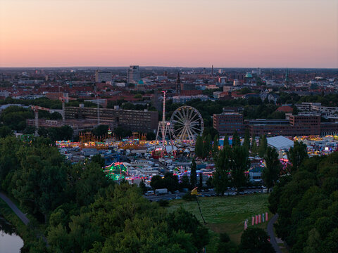 Hanover, Lower Saxony, Germany - 12 July 2026: Aerial view of Maschsee fairground with a Ferris wheel and illuminated carnival rides at sunset, with the city skyline of Hanover in the background.