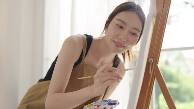 Creative young Asian woman artist painting on canvas in bright home studio. Relaxed female hobbyist enjoying peaceful indoor leisure time, expressing imagination and passion for art.