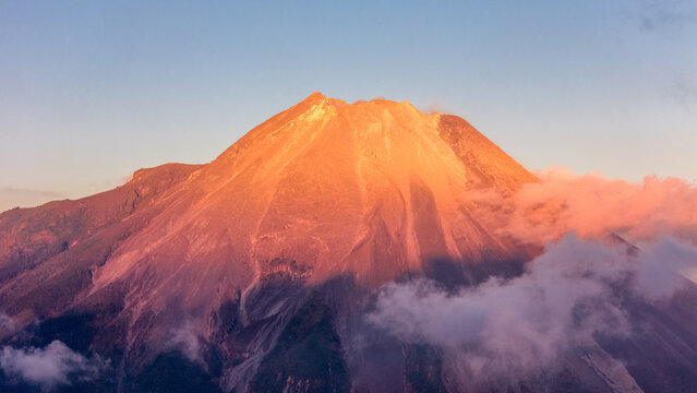 Aerial view of Mount Merapi illuminated by golden hour light with clouds drifting around its slopes in Kecamatan Dukun, Jawa Tengah, Indonesia.