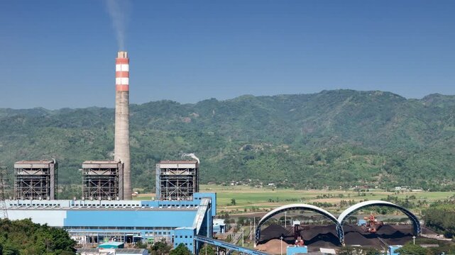 Aerial view of industrial power plant facility with smoking chimney and coal storage domes set against green mountains in Indonesia.