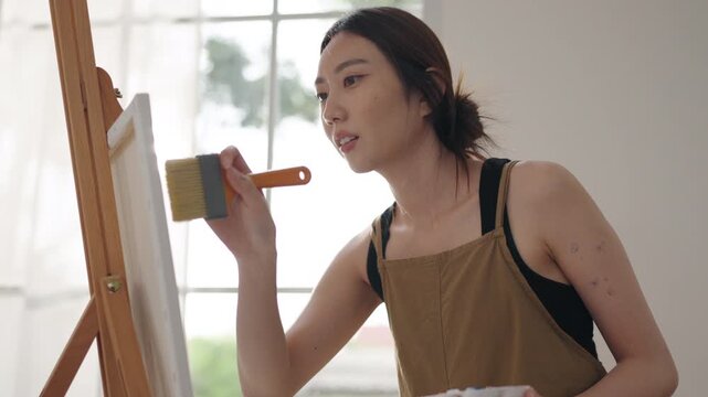 Creative young Asian woman artist painting on canvas in bright home studio. Relaxed female hobbyist sitting on floor, enjoying peaceful indoor leisure time and expressing imagination.