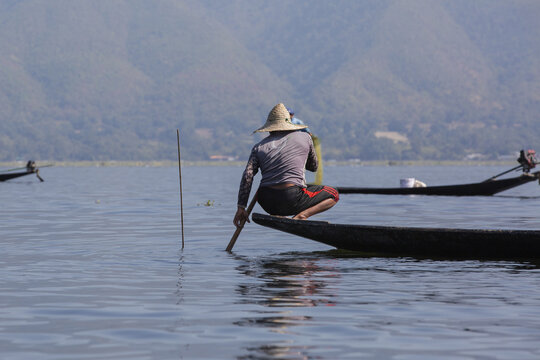 View of a fisherman wearing a conical hat balancing on a traditional wooden boat on Inle Lake with mountains in the background in Taunggyi, Shan, Myanmar.