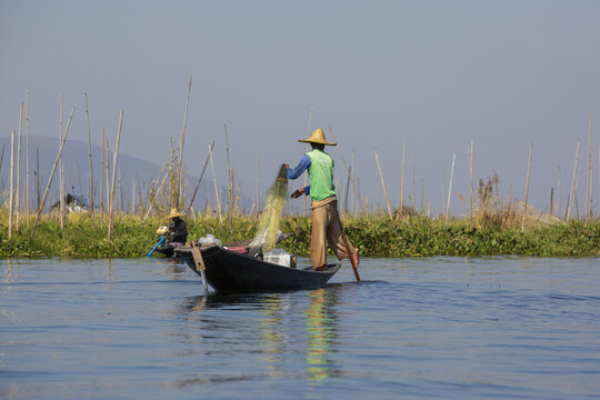 View of a fisherman casting a net from a traditional wooden boat on Inle Lake with mountains in the background in Taunggyi, Shan, Myanmar.
