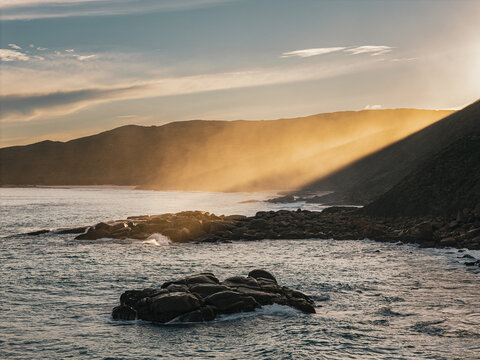 Aerial view of the Blowholes at Torndirrup National Park with golden hour light hitting the rugged coastline, dark rocky outcrops, and crashing waves in Torndirrup, Western Australia, Australia.