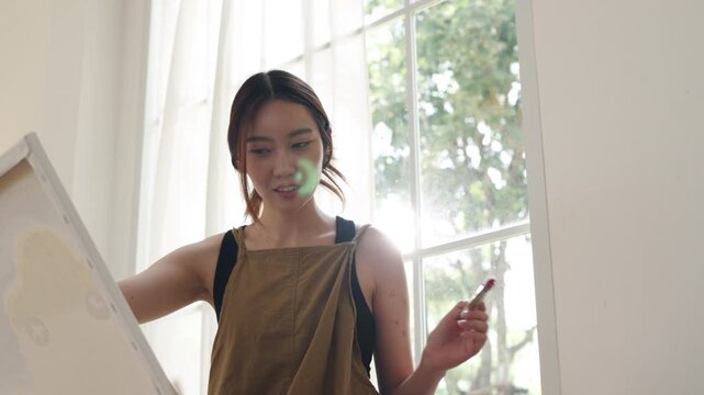 Creative young Asian woman artist painting on canvas in bright home studio. Relaxed female hobbyist sitting on floor, enjoying peaceful indoor leisure time and expressing imagination.