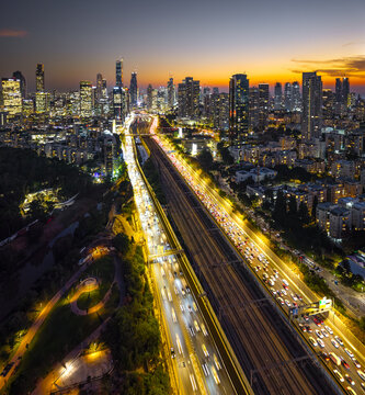 Aerial view of the illuminated Tel Aviv skyline at night with vibrant light trails from heavy traffic on the highway and a glowing sunset sky in Tel Aviv-Yafo, Tel Aviv District, Israel.