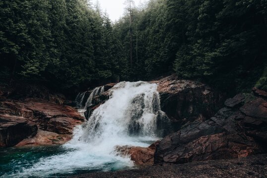 View of a cascading waterfall surrounded by dark mossy rocks and a dense evergreen forest in Vancouver, British Columbia, Canada.