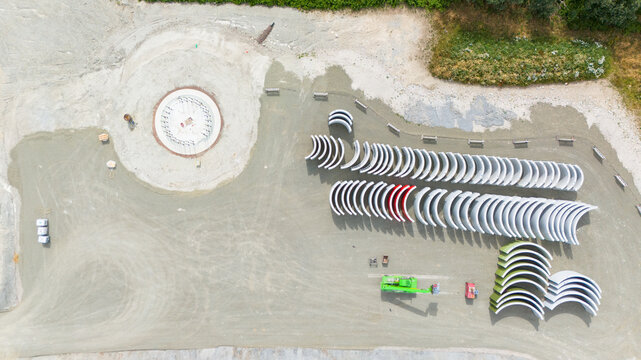 Aerial view of a wind turbine construction site featuring a circular foundation and rows of curved concrete tower segments on a gravel ground in Trier-Saarburg, Rhineland-Palatinate, Germany.