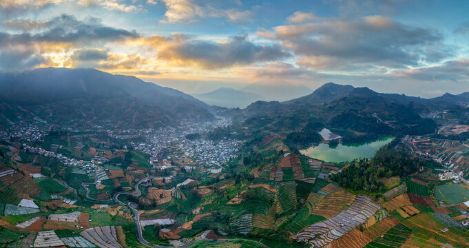 Aerial view of Telaga Warna lake surrounded by lush agricultural terraces, rolling hills, and the village of Kejajar under a golden sunset sky in Kejajar, Central Java, Indonesia.