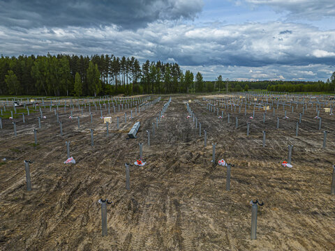 Aerial view of a construction site for a solar farm with metal support posts installed in the ground, surrounded by a forest under a cloudy sky in Riga, Latvia.