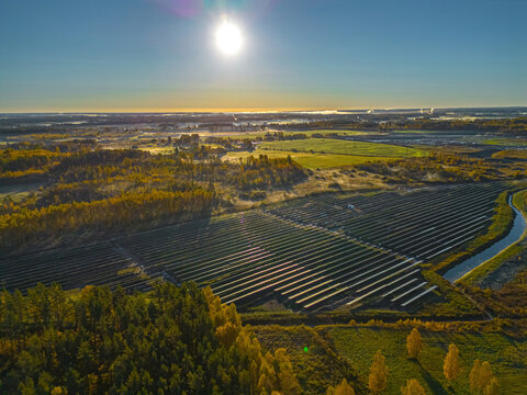 Aerial view of a large solar farm with rows of photovoltaic panels set in a rural landscape with autumn trees and a small stream under a bright sun in Riga, Latvia.