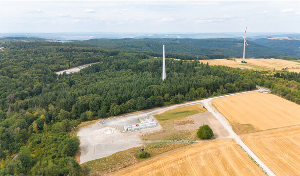 Aerial view of wind turbine construction site in a forested area with cleared land, turbine components, and a completed turbine in the distance, Trier-Saarburg, Rhineland-Palatinate, Germany.