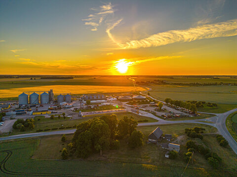 Aerial view of an agricultural facility with grain silos and farm buildings surrounded by vast fields during a golden sunset in Dobele Municipality, Latvia.