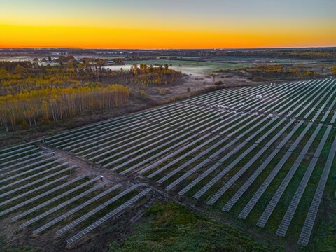 Aerial view of a large solar farm with rows of photovoltaic panels set against a vibrant sunset sky over the rural landscape in Riga, Latvia.