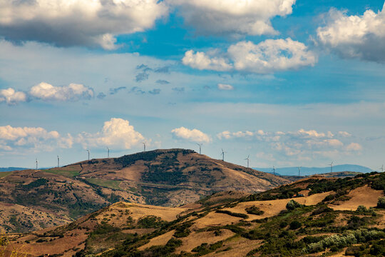 View of wind turbines on a rolling hill ridge surrounded by dry Mediterranean landscape under a blue sky in Sardinia, Italy.