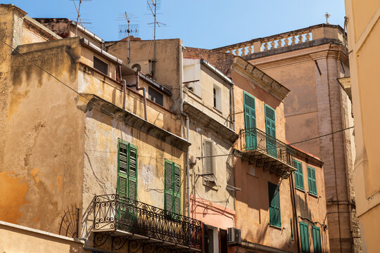 View of historic residential buildings with weathered facades, green shutters, and ornate wrought iron balconies in Sardinia, Italy.