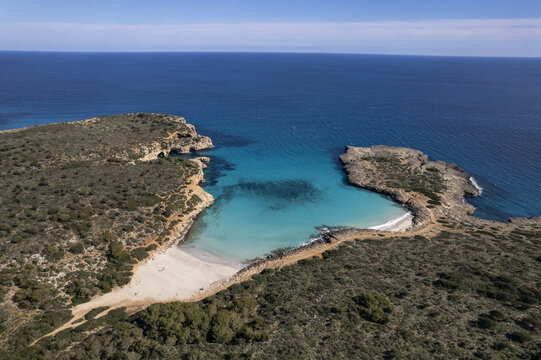 Aerial view of Cala Varques with its turquoise waters, sandy beach, and rocky cliffs surrounded by Mediterranean vegetation, Mallorca, Spain.