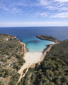 Aerial view of Cala Varques featuring turquoise waters, a pristine sandy beach, and rugged rocky coastline surrounded by Mediterranean vegetation in Mallorca, Spain.