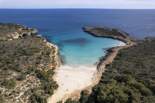 Aerial view of Cala Varques with its turquoise waters, sandy beach, and surrounding rocky cliffs covered in Mediterranean vegetation, Mallorca, Spain.