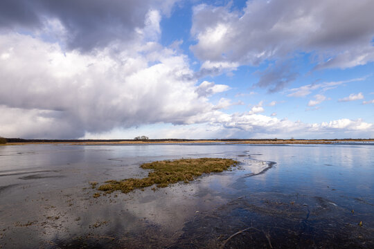 View of Biebrza wetlands with a small grassy island in a partially frozen marsh under a dramatic cloudy sky in Podlaskie Voivodeship, Poland.