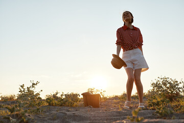 Obraz premium Happy woman enjoying lifestyle during summer picnic on sandy beach at sunset. She holds straw hat and smiles, wearing casual clothes. Concept of freedom, joy, and relaxation.