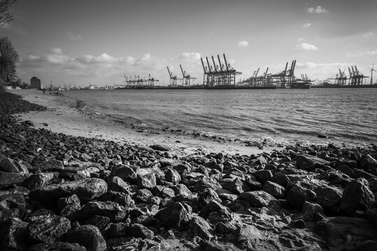 Hamburg, Germany - 14 July 2026: View of the Port of Hamburg with large container cranes lining the riverbank under a cloudy sky.