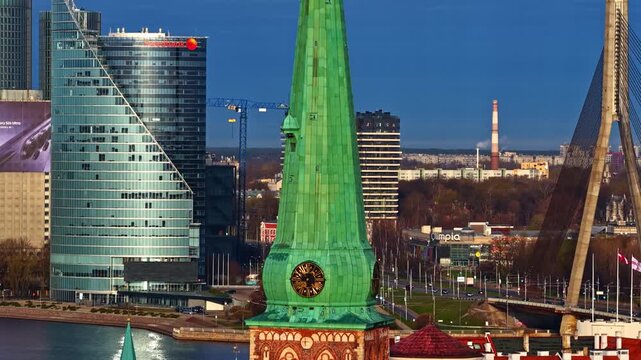 Aerial view of the historic St. Peter's Church spire alongside modern skyscrapers and the Vansu Bridge over the Daugava River in Riga, Latvia.
