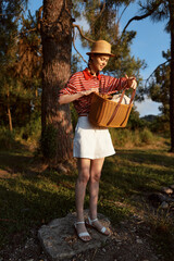 Obraz premium Young woman enjoying a lifestyle picnic outdoors. She holds a woven basket while standing near a tree in the forest. A sunny summer day concept with a beautiful nature setting.