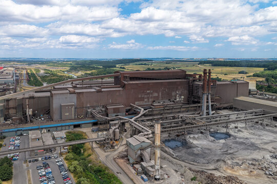 Aerial view of a large industrial steel mill complex with tall smokestacks, complex piping systems, and a parking lot, surrounded by green fields and a blue sky in the United Kingdom.