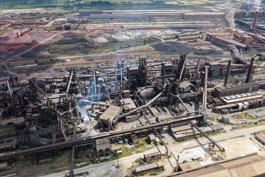 Aerial view of a massive industrial steel plant featuring complex blast furnaces, intricate piping networks, and smoke stacks emitting plumes in the United Kingdom.