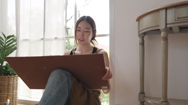Creative young Asian woman artist painting on canvas in bright home studio. Relaxed female hobbyist sitting on floor, enjoying peaceful indoor leisure time and expressing imagination.