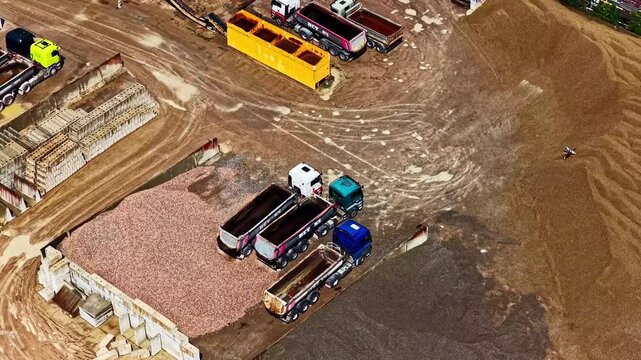 Aerial view of a construction site featuring several dump trucks parked near large piles of gravel and sand in an industrial area in Jelgava, Jelgava City, Latvia.