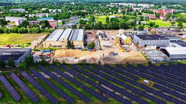 Aerial view of a solar farm with rows of photovoltaic panels near industrial buildings and green fields in Jelgava, Jelgava pilseta, Latvia.
