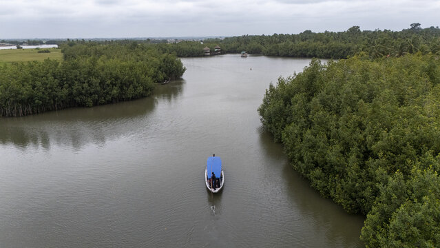 Aerial view of a small boat with a blue canopy navigating the calm waters of the Gogotinkpon lagoon, surrounded by dense mangrove forests under a cloudy sky in Kpomasse, Mono, Benin.