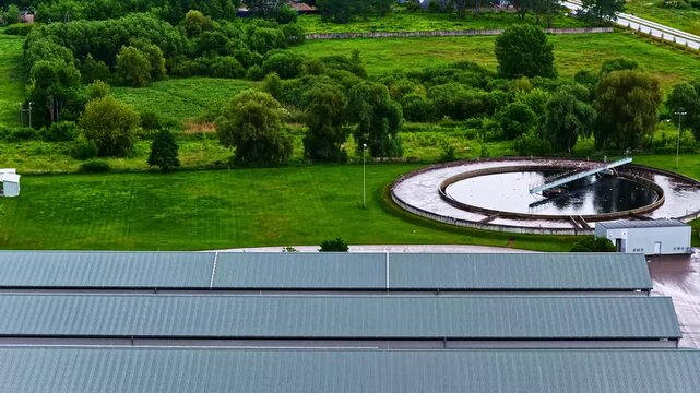 Aerial view of a wastewater treatment plant featuring a large circular sedimentation tank surrounded by green fields and industrial buildings in Jelgava, Jelgava pilseta, Latvia.