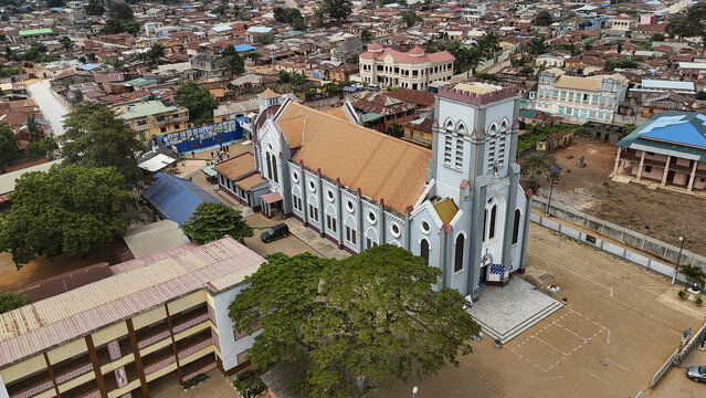 Aerial view of the Basilica of the Immaculate Conception surrounded by historic urban rooftops, lush trees, and a courtyard in Ouidah, Atlantique, Benin.