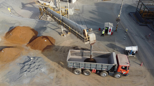 Aerial view of a mining site with a conveyor belt loading raw material into a dump truck, piles of ore, and a worker walking on the ground in Barranca, Lima Region, Peru.
