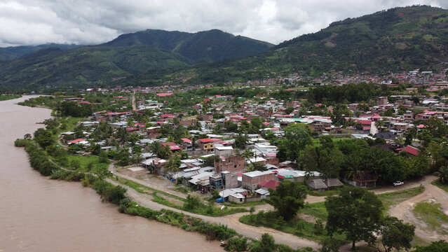 Aerial view of Pichanaki town nestled along the riverbank with lush green mountains in the background, featuring dense residential buildings and tropical vegetation in Pichanaki, Junin, Peru.
