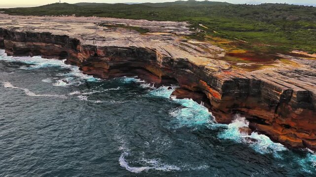 Aerial view of rugged sandstone cliffs along the coastline with crashing waves and coastal vegetation in Kurnell, New South Wales, Australia.
