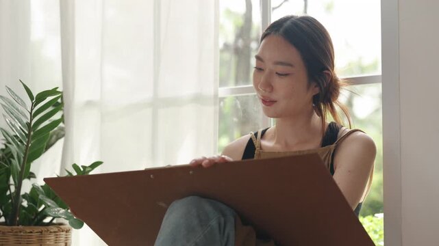 Creative young Asian woman artist painting on canvas in bright home studio. Relaxed female hobbyist sitting on floor, enjoying peaceful indoor leisure time and expressing imagination.