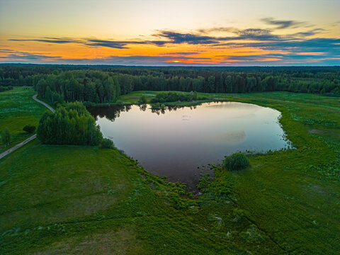 Aerial view of a serene lake surrounded by lush green fields and dense forests during a vibrant golden hour sunset in Vidzeme, Latvia.