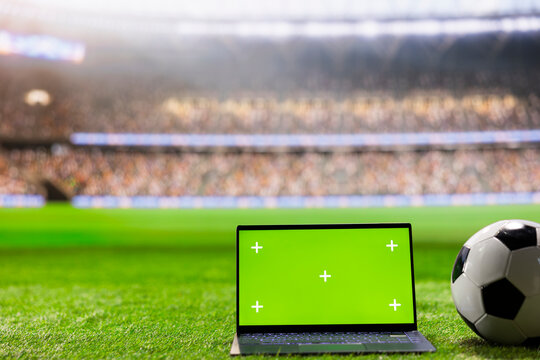 Laptop green screen mockup sits on stadium grass beside a soccer ball, offering screen replacement space for digital sports content against blurred arena stands and crowd on a football pitch.