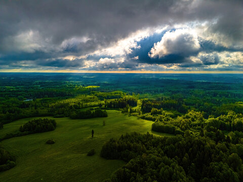 Aerial view of rolling green hills and dense forests under dramatic clouds with sunlight breaking through in Vidzeme, Latvia.
