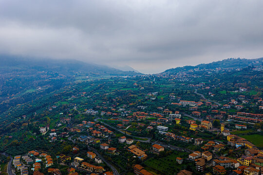 Aerial view of Lake Como surrounded by lush green hills, scattered residential buildings with terracotta roofs, and winding roads under a cloudy sky in Lombardy, Italy.