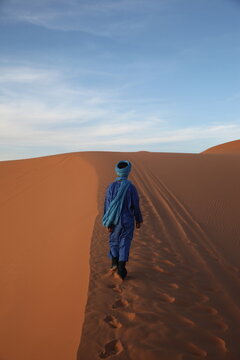View of a person wearing a blue djellaba and turban walking across the vast orange sand dunes of the Sahara desert in Morocco.