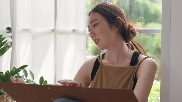 Creative young Asian woman artist painting on canvas in bright home studio. Relaxed female hobbyist sitting on floor, enjoying peaceful indoor leisure time and expressing imagination.