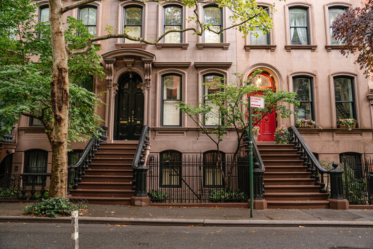 Traditional New York brownstone townhouses with symmetrical staircases, detailed doors, and large windows along a quiet street. Historic brick residential architecture surrounded by trees in a