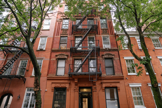 Upward view of a classic New York brownstone apartment building with black metal fire escape and red brick facade framed by green trees. Urban Manhattan architecture featuring detailed windows