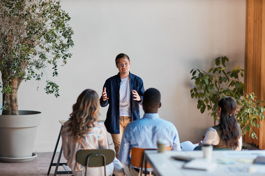 Young male speaker giving a business presentation during a team meeting in a modern office, sharing ideas and expertise with diverse colleagues for professional development