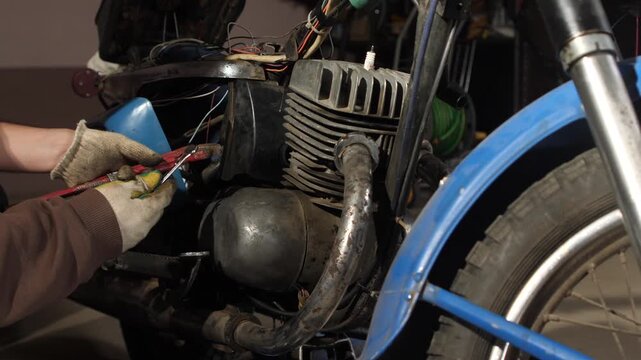 Mechanic's hands in dirty work gloves using a red pipe wrench to perform maintenance and repair work on the engine of a vintage blue motorcycle in a garage